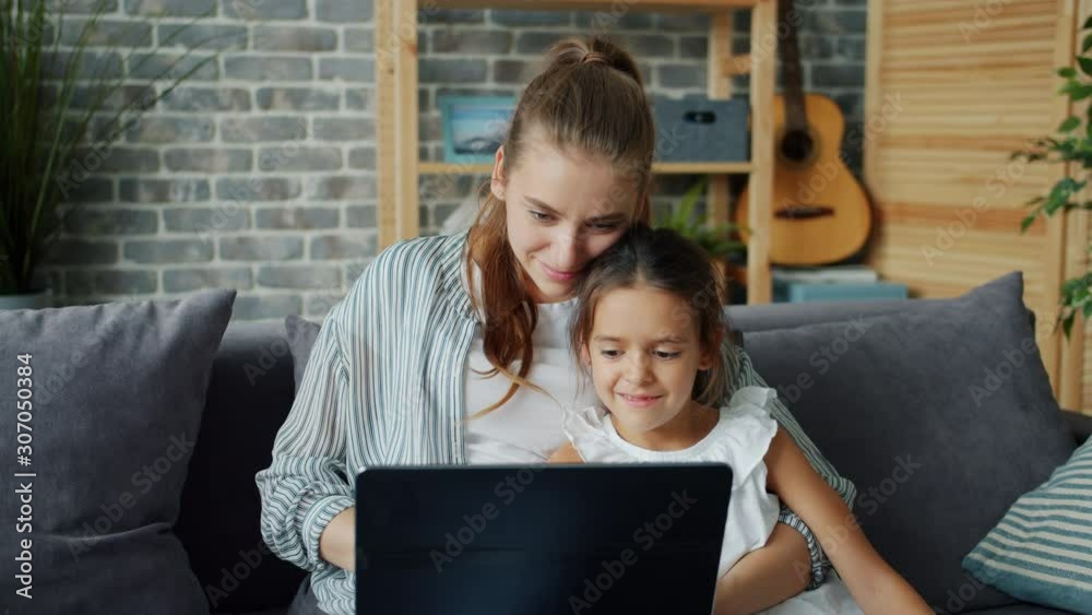 Mother and little daughter are using laptop looking at screen enjoying modern technology at home smiling together. Modern devices, family and entertainment concept.