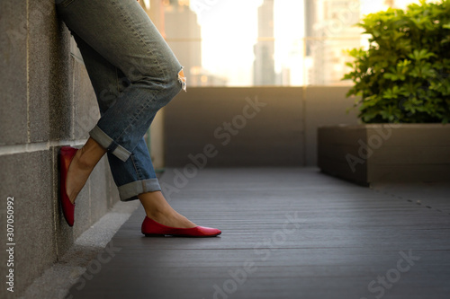 Crop shot of an attractive woman's legs in smart casual outfit, jeans and ruby red flat shoes, she stand and lean against the wall on building rooftop in the evening. Modern city leisure lifestyle.