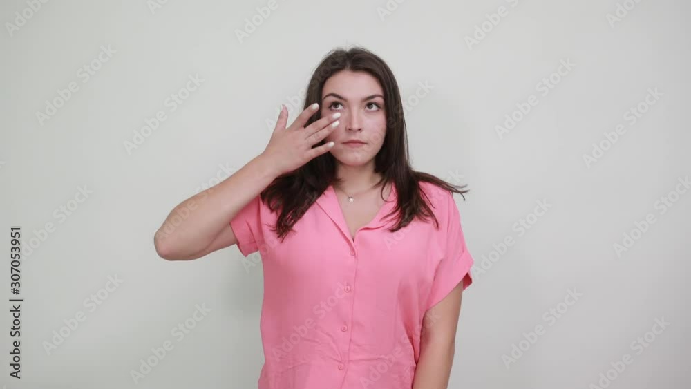 Serious young woman keeping fingers under eyes, wipes away tears isolated on gray background in studio in casual pink shirt. People sincere emotions, lifestyle concept. Serious young woman keeping fingers under eyes, wipes away tears isolated on gray background in studio in casual pink shirt. People sincere emotions, lifestyle concept.