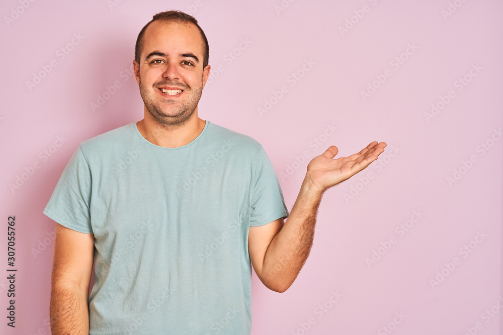 Young man wearing blue casual t-shirt standing over isolated pink background smiling cheerful presenting and pointing with palm of hand looking at the camera. Young man wearing blue casual t-shirt standing over isolated pink background smiling cheerful presenting and pointing with palm of hand looking at the camera.