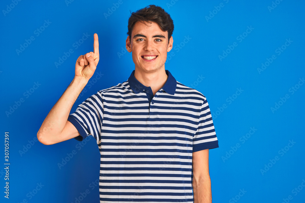 Teenager boy wearing casual t-shirt standing over blue isolated background showing and pointing up with finger number one while smiling confident and happy.