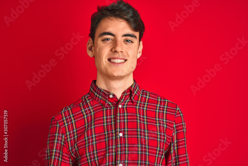 Teenager boy wearing red shirt standing over isolated background looking away to side with smile on face, natural expression. Laughing confident.