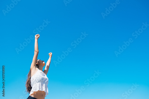 Happy woman with tanned slim body breathing fresh air raising her arms up, enjoying a sunny summer holiday on beach destination against blue sky, outdoors. Travel and well being lifestyle.