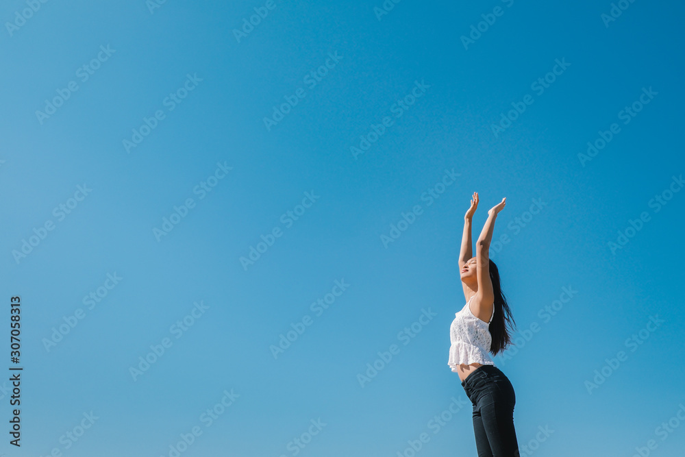 Happy woman with tanned slim body breathing fresh air raising her arms up, enjoying a sunny summer holiday on beach destination against blue sky, outdoors. Travel and well being lifestyle.