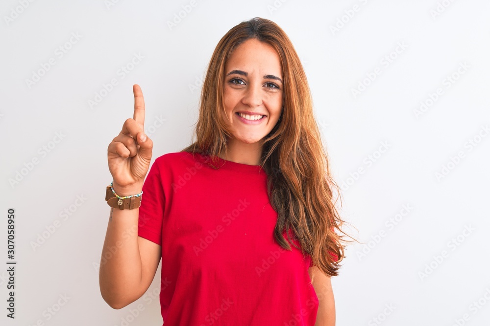 Young redhead woman wearing red casual t-shirt stading over white isolated background showing and pointing up with finger number one while smiling confident and happy. Young redhead woman wearing red casual t-shirt stading over white isolated background showing and pointing up with finger number one while smiling confident and happy.