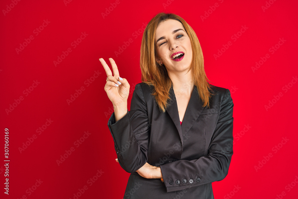 Young caucasian business woman wearing a suit over isolated red background smiling with happy face winking at the camera doing victory sign. Number two. Young caucasian business woman wearing a suit over isolated red background smiling with happy face winking at the camera doing victory sign. Number two.