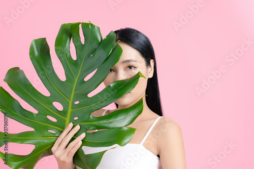 Asian woman holding a tropical leaf with natural skin on pink background. Beauty concept.