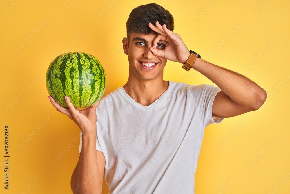 Young indian shopkeeper man holding watermelon standing over isolated yellow background with happy face smiling doing ok sign with hand on eye looking through fingers