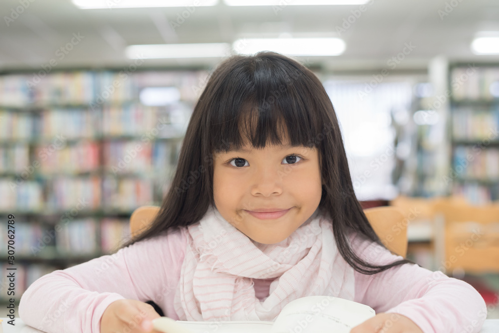 portrait of cute schoolgirl smiling while sitting with stack of books at table in library