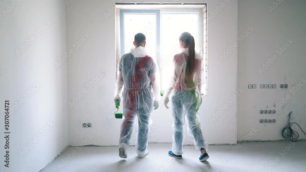Dolly cam: young blonde woman and handsome man wearing transparent coveralls come to window joining hands in empty room Dolly cam: young blonde woman and handsome man wearing transparent coveralls come to window joining hands in empty room