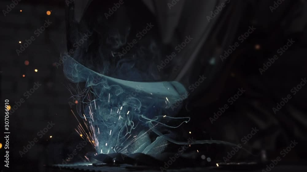 Forge workshop. Smithy. Worker in a welding hood helmet welds a part by electric welding. Sparks are reflected in the protective screen. Blacksmith makes iron product for manufacture. Slow motion.