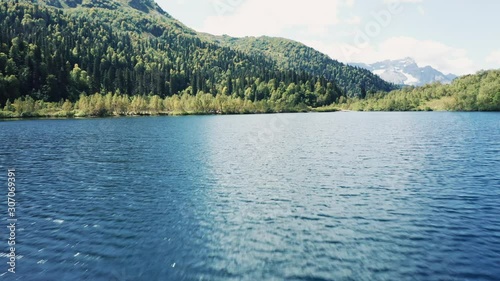 Aerial view; drone flying low over emerald water surfase of lake surrounded by mountain ranges and valleys; reflecting of lakesides on water; Agepsta mountain range with glaciers on background