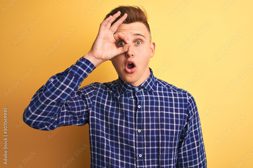 Young handsome man wearing casual shirt standing over isolated yellow background doing ok gesture shocked with surprised face, eye looking through fingers. Unbelieving expression.