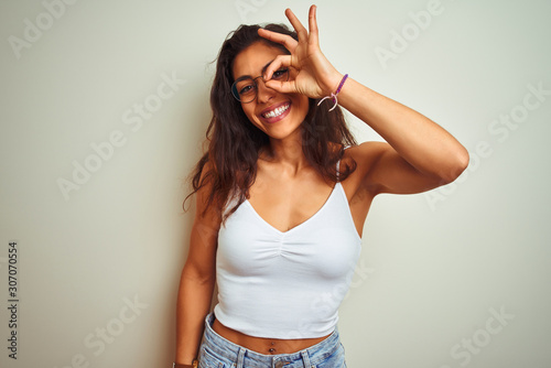 Young beautiful woman wearing t-shirt and glasses standing over isolated white background doing ok gesture with hand smiling, eye looking through fingers with happy face.