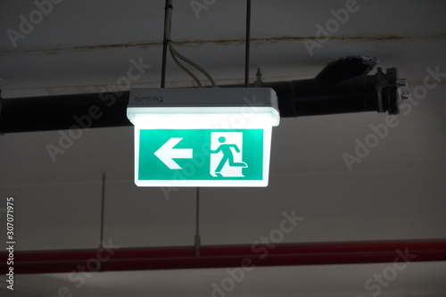 Glowing Emergency exit sign with left arrow at a building. Safety first concept. Copy space wallpaper. Exit signs with light in a parking of building.