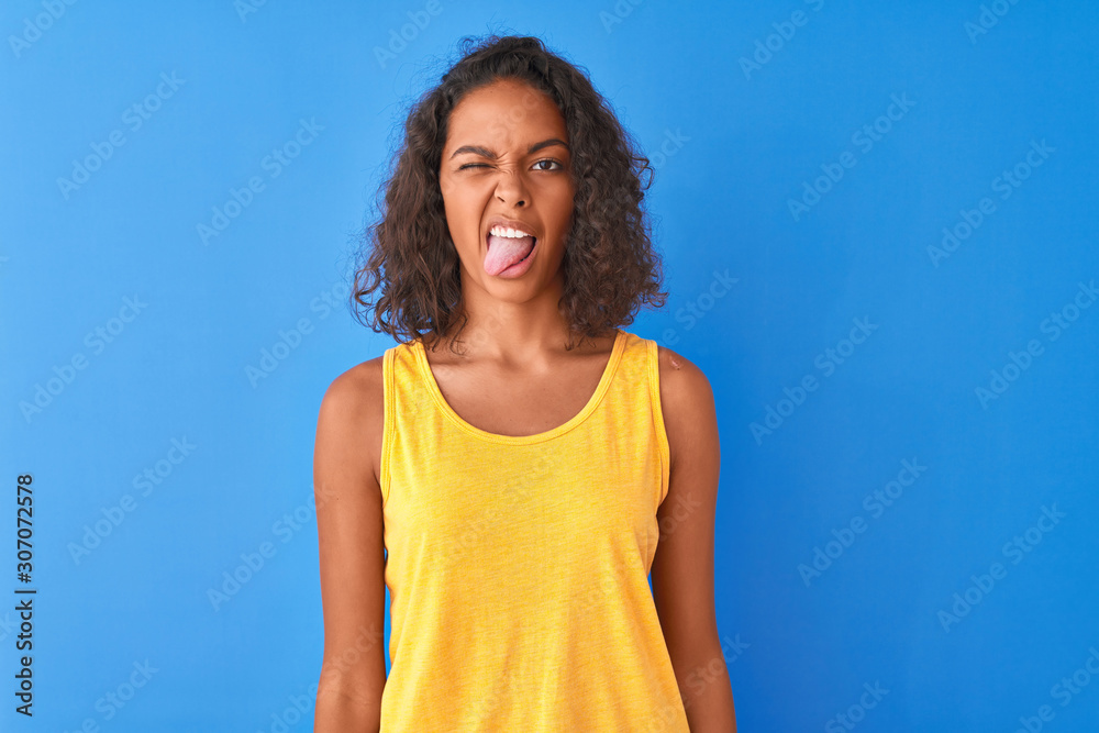 Young brazilian woman wearing yellow t-shirt standing over isolated blue background sticking tongue out happy with funny expression. Emotion concept. Young brazilian woman wearing yellow t-shirt standing over isolated blue background sticking tongue out happy with funny expression. Emotion concept.