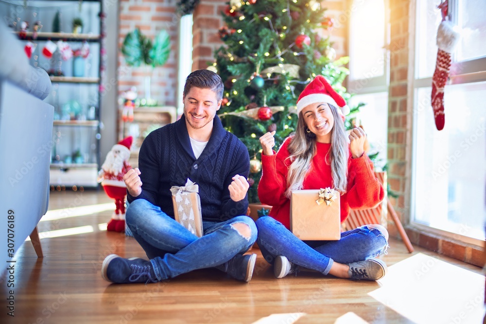 Young couple wearing santa claus hat sitting on the floor around christmas tree at home very happy and excited doing winner gesture with arms raised, smiling and screaming for success. Celebration Young couple wearing santa claus hat sitting on the floor around christmas tree at home very happy and excited doing winner gesture with arms raised, smiling and screaming for success. Celebration