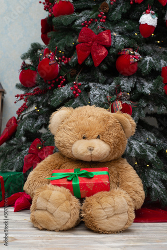 Toy teddy bear with a Christmas present sits by the Christmas tree