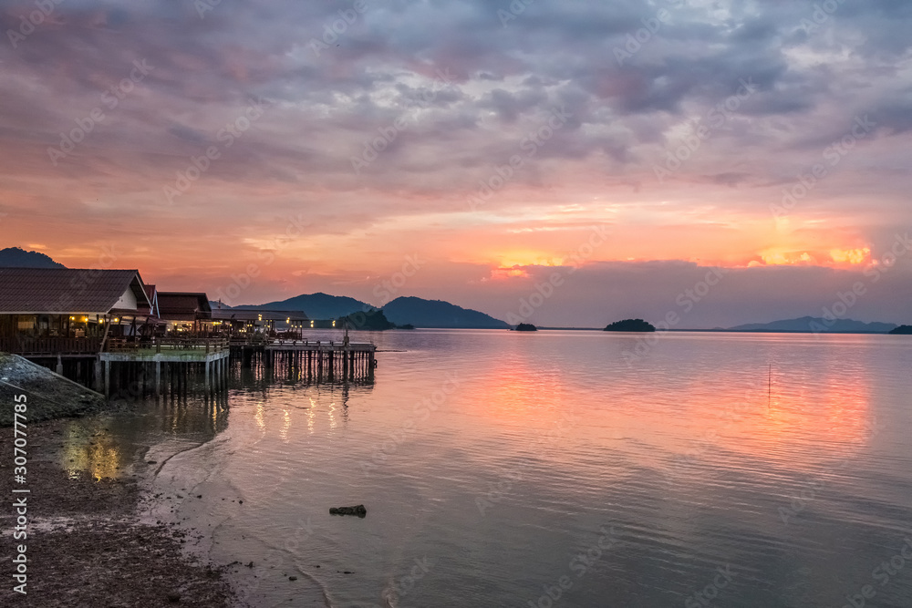 Beautiful sunset in Thailand on Koh Lanta. Along the shore are houses with evening illumination. On the horizon are mountain hills. The looming cloudy sky is reflected in the calm sea