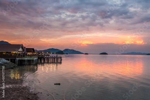 Beautiful sunset in Thailand on Koh Lanta. Along the shore are houses with evening illumination. On the horizon are mountain hills. The looming cloudy sky is reflected in the calm sea
