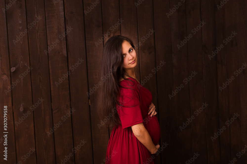 Fototapeta premium Pregnant woman in red dress holds hands on belly on a dark brown background. Pregnancy, maternity, expectation concept. Beautiful tender mood photo of pregnancy