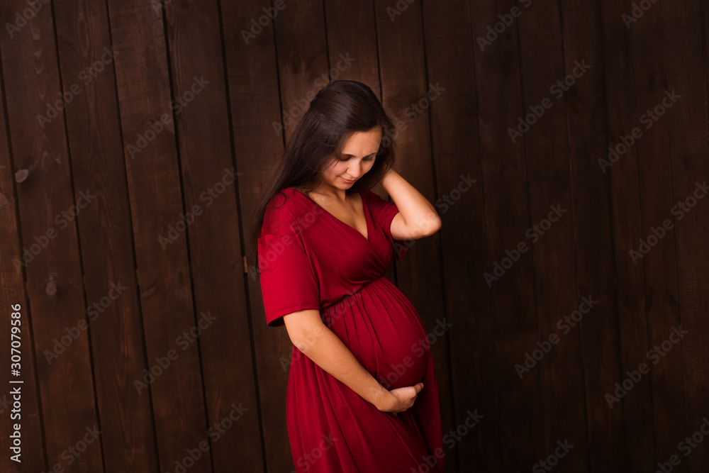 Fototapeta premium Pregnant woman in red dress holds hands on belly on a dark brown background. Pregnancy, maternity, expectation concept. Beautiful tender mood photo of pregnancy