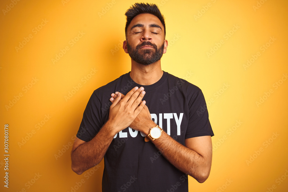 Arab indian hispanic safeguard man wearing security uniform over isolated yellow background smiling with hands on chest with closed eyes and grateful gesture on face. Health concept. Arab indian hispanic safeguard man wearing security uniform over isolated yellow background smiling with hands on chest with closed eyes and grateful gesture on face. Health concept.