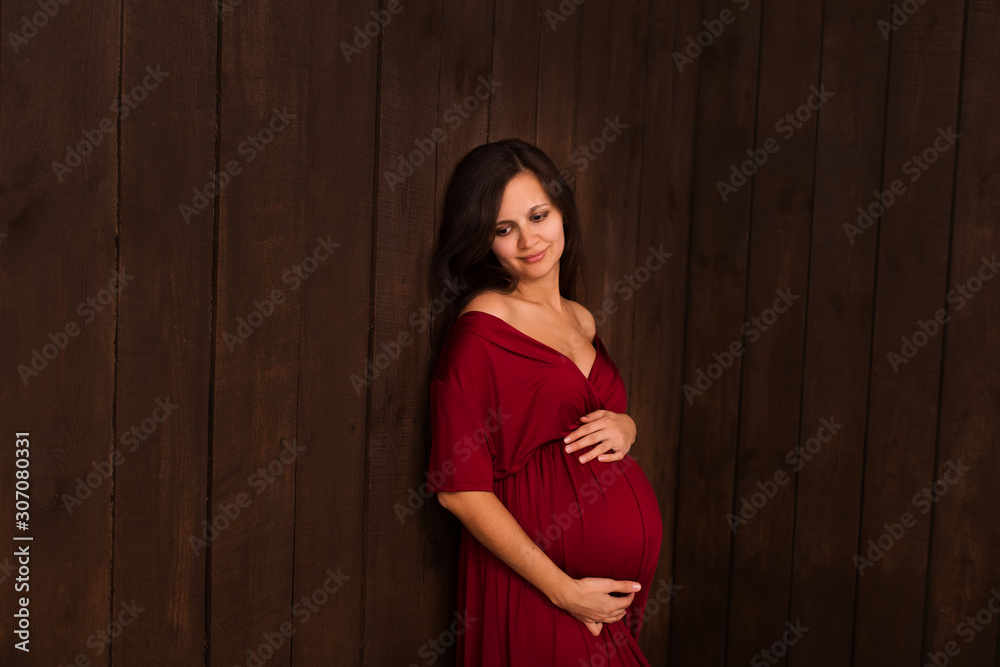 Fototapeta premium Christmas pregnant woman in red dress holds hands on belly on a dark brown background. Pregnancy, maternity, expectation concept. Beautiful tender mood photo of pregnancy