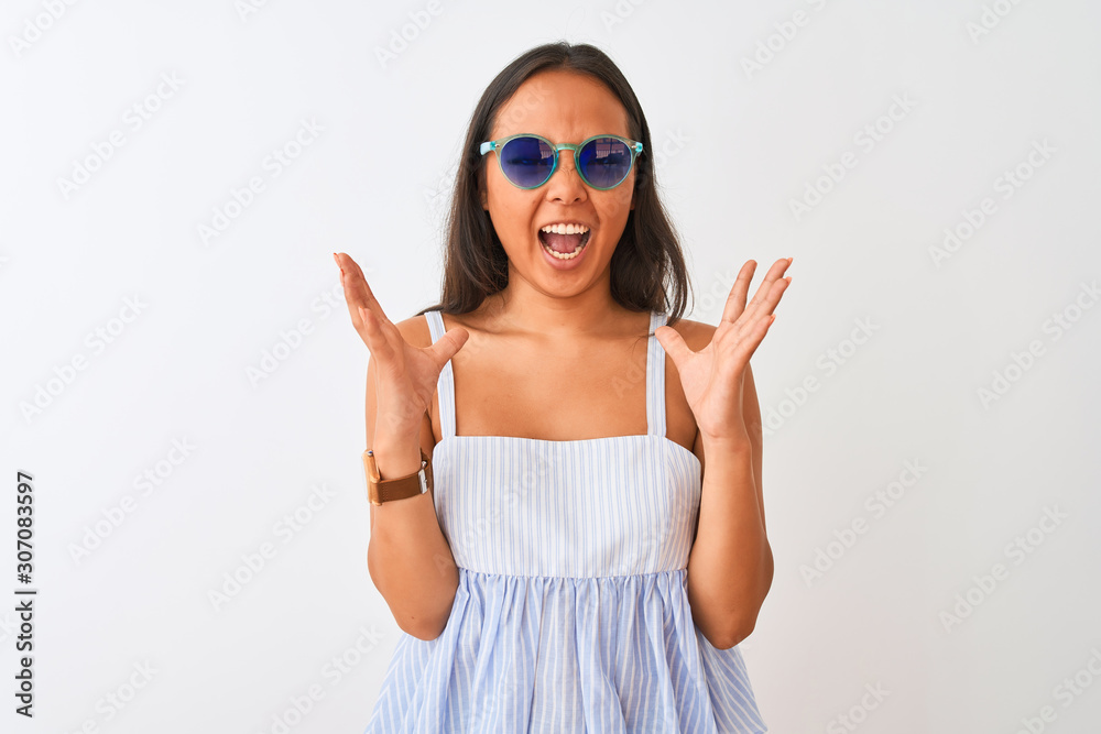 Young chinese woman wearing striped dress and sunglasses over isolated white background celebrating mad and crazy for success with arms raised and closed eyes screaming excited. Winner concept Young chinese woman wearing striped dress and sunglasses over isolated white background celebrating mad and crazy for success with arms raised and closed eyes screaming excited. Winner concept