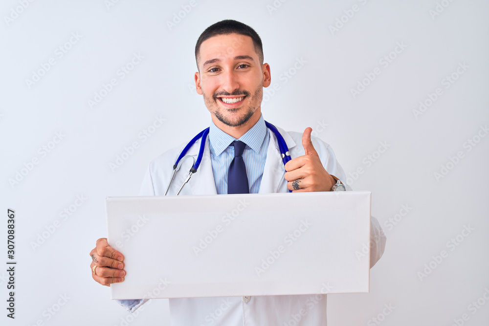 Young doctor man holding blank advertising banner over isolated background happy with big smile doing ok sign, thumb up with fingers, excellent sign Young doctor man holding blank advertising banner over isolated background happy with big smile doing ok sign, thumb up with fingers, excellent sign