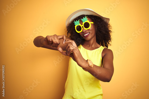African american woman wearing funny pineapple sunglasses over isolated yellow background smiling in love showing heart symbol and shape with hands. Romantic concept.