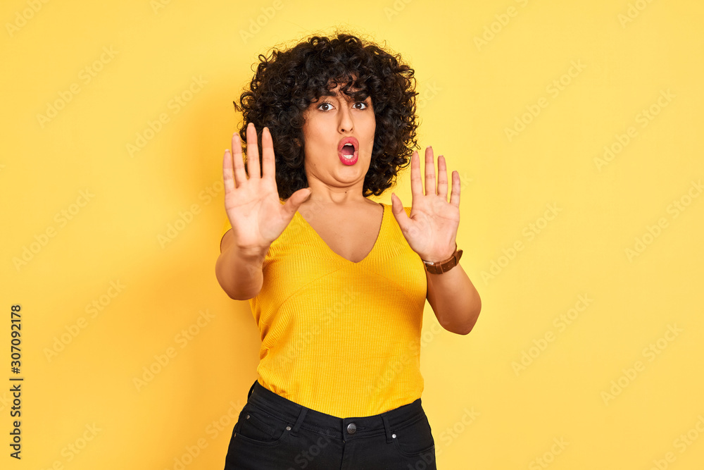 Young arab woman with curly hair wearing t-shirt standing over isolated yellow background afraid and terrified with fear expression stop gesture with hands, shouting in shock. Panic concept.
