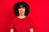 Young arab woman with curly hair wearing casual t-shirt over isolated red background sticking tongue out happy with funny expression. Emotion concept.