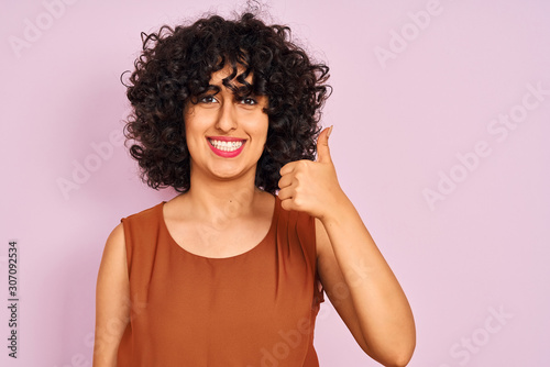 Young arab woman with curly hair wearing t-shirt over isolated pink background happy with big smile doing ok sign, thumb up with fingers, excellent sign