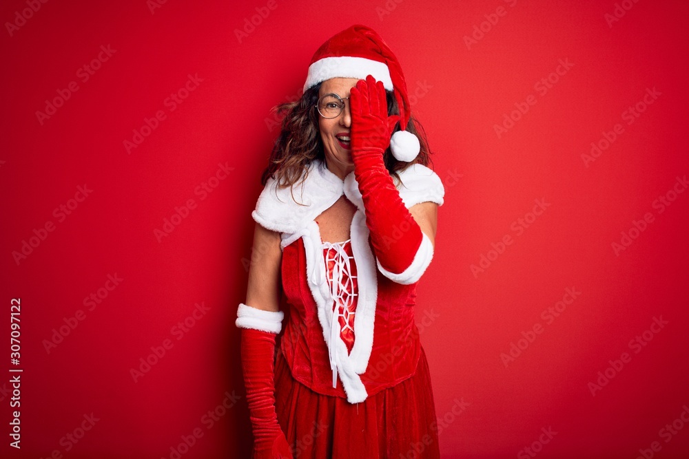 Middle age beautiful woman wearing Santa Claus costume over isolated red background covering one eye with hand, confident smile on face and surprise emotion. Middle age beautiful woman wearing Santa Claus costume over isolated red background covering one eye with hand, confident smile on face and surprise emotion.