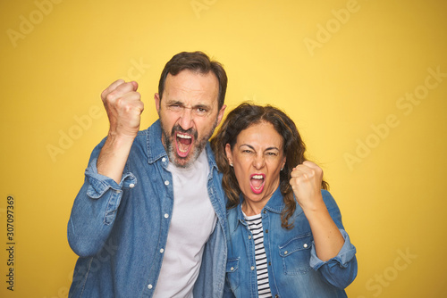 Beautiful middle age couple together wearing denim shirt over isolated yellow background angry and mad raising fist frustrated and furious while shouting with anger. Rage and aggressive concept.