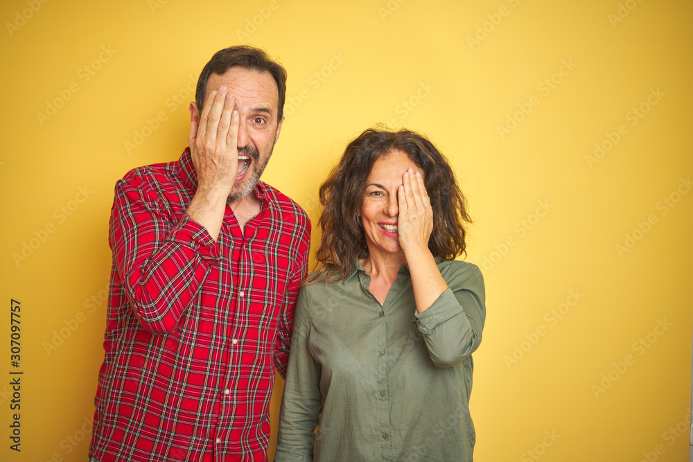 Beautiful middle age couple over isolated yellow background covering one eye with hand, confident smile on face and surprise emotion. Beautiful middle age couple over isolated yellow background covering one eye with hand, confident smile on face and surprise emotion.