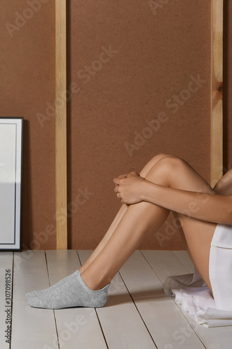 Cropped photo of a girl's lower body's part, sitting on a floor. There are short grey socks on her feet. The photo was taken against the background of a brown wall and a white wooden floor. photo