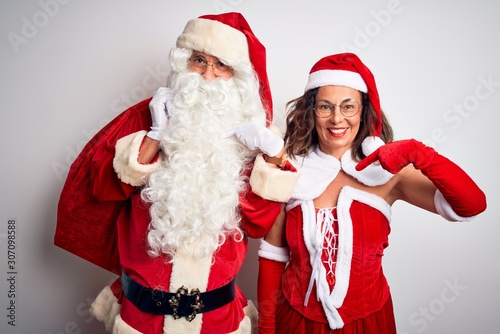 Senior couple wearing Santa Claus costume holding sack over isolated white background with surprise face pointing finger to himself