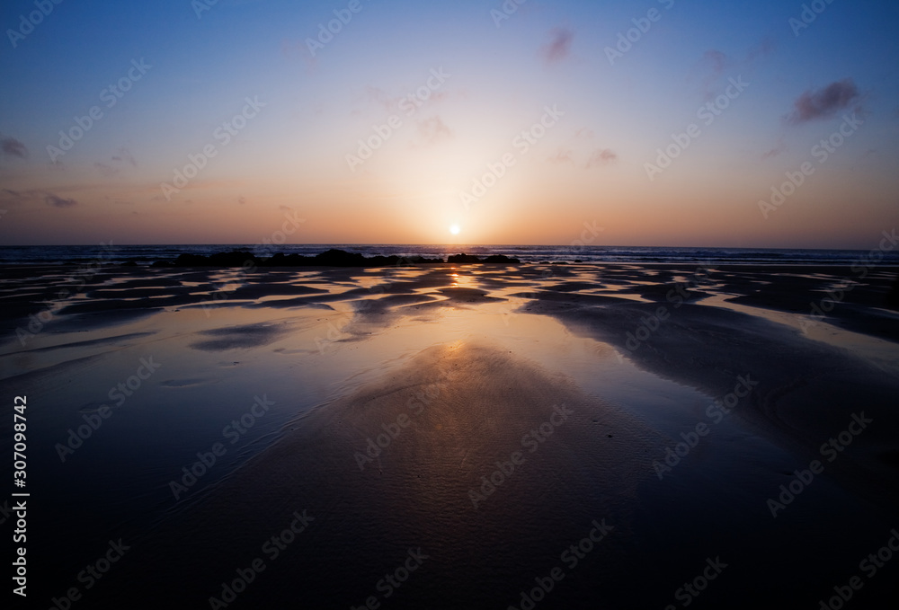 Sun sinks behind horizon at Porthtowan, Cornwall with colourful reflections in the pools of water on the beach.