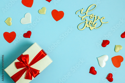 Valentines day background copy space. On a blue background a gift box and a red bow, red paper hearts, white fabric and red satin hearts and the inscription made of wooden letters, I love you