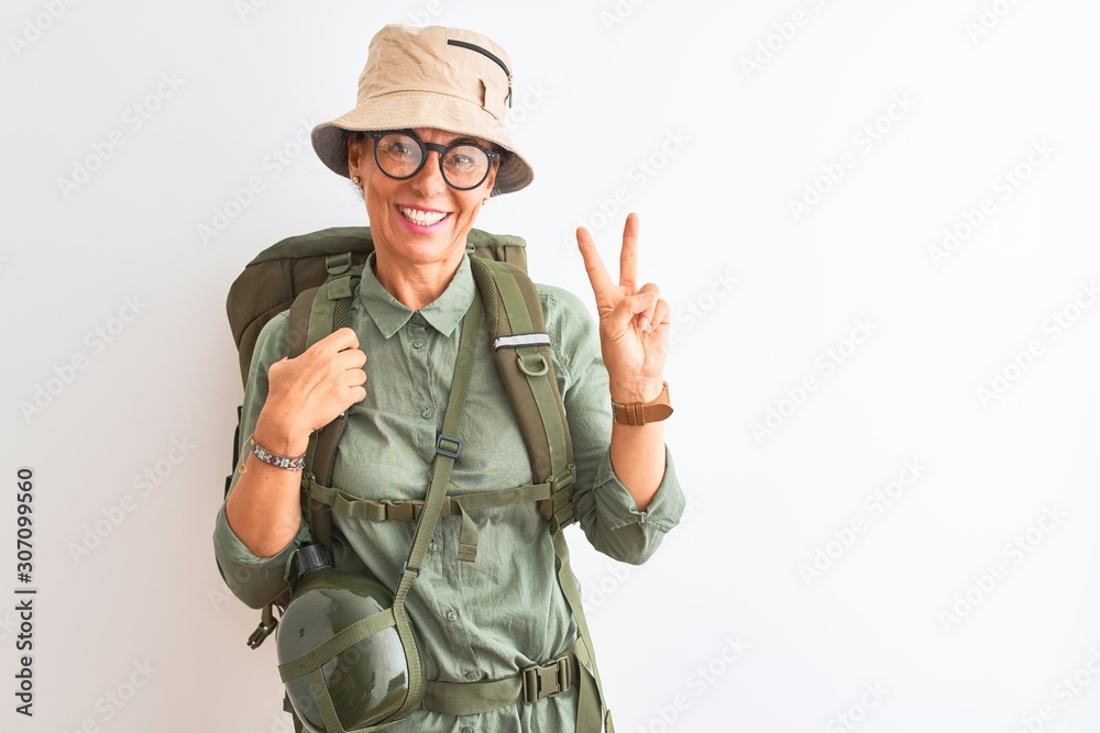 Fototapeta premium Middle age hiker woman wearing backpack canteen hat glasses over isolated white background smiling with happy face winking at the camera doing victory sign. Number two.