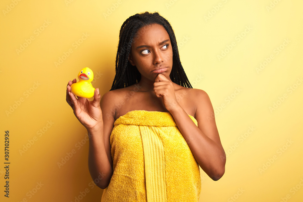 African american woman wearing shower towel holding toy duck over isolated yellow background serious face thinking about question, very confused idea African american woman wearing shower towel holding toy duck over isolated yellow background serious face thinking about question, very confused idea