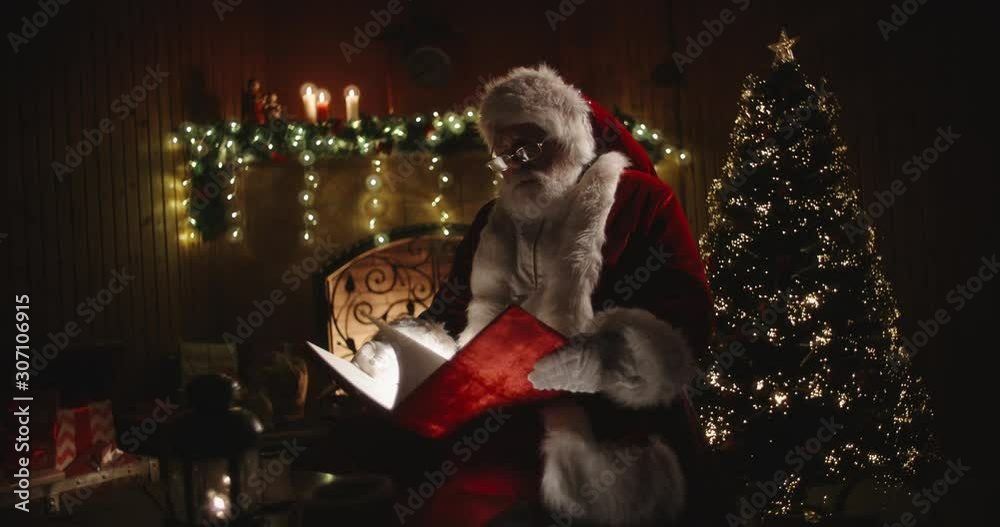 Authentic santa clause opening his magical shining book with red cover, sitting on rocker near fireplace and christmas tree - holiday mood, christmas spirit concept 4k footage