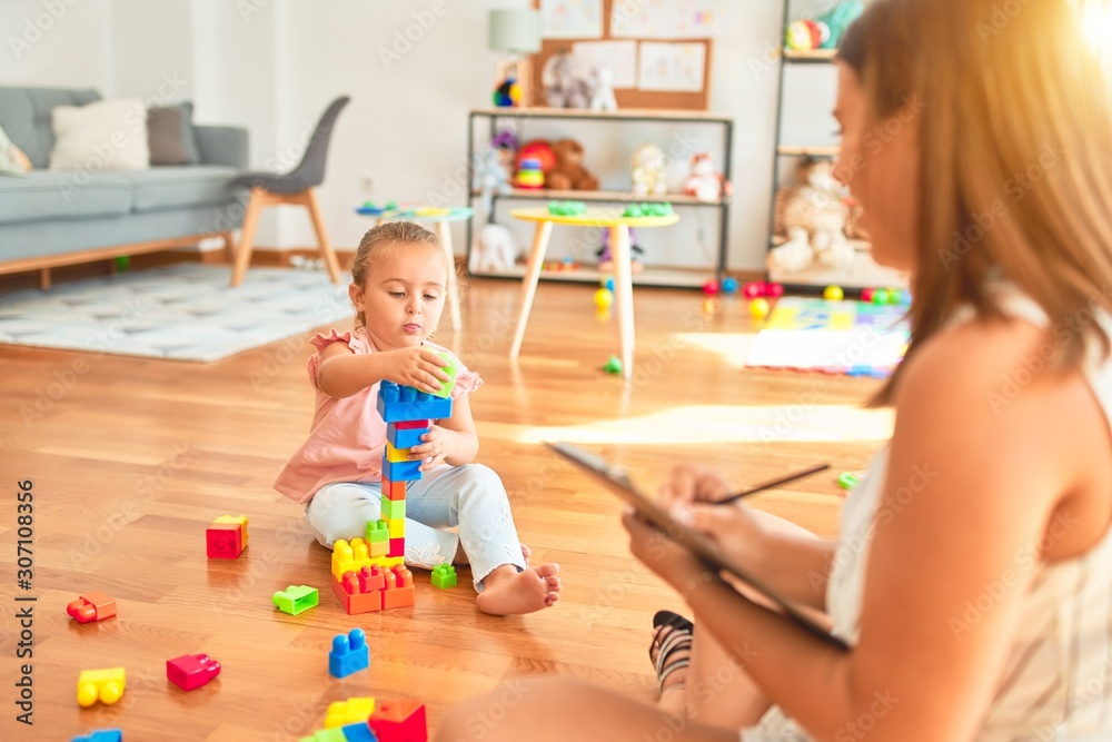 Beautiful psycologist and blond toddler girl doing therapy building tower using plastic blocks at kindergarten Beautiful psycologist and blond toddler girl doing therapy building tower using plastic blocks at kindergarten