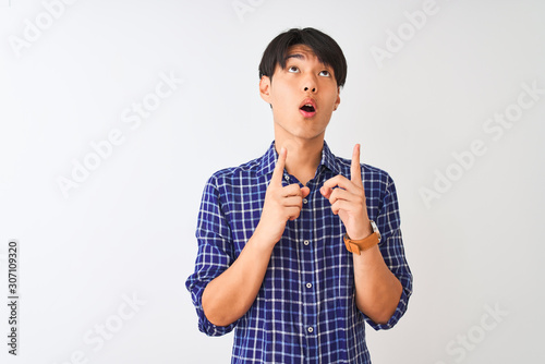 Young chinese man wearing casual blue shirt standing over isolated white background amazed and surprised looking up and pointing with fingers and raised arms.