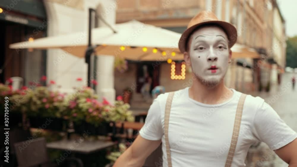 Young handsome mime is performing on street near cafe, denies, says no certainly. Performing by entertainer in hat, suspenders, makeup. Artist is moving arms, looks scared.
