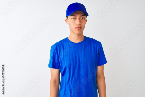 Chinese deliveryman wearing blue t-shirt and cap standing over isolated white background Relaxed with serious expression on face. Simple and natural looking at the camera.