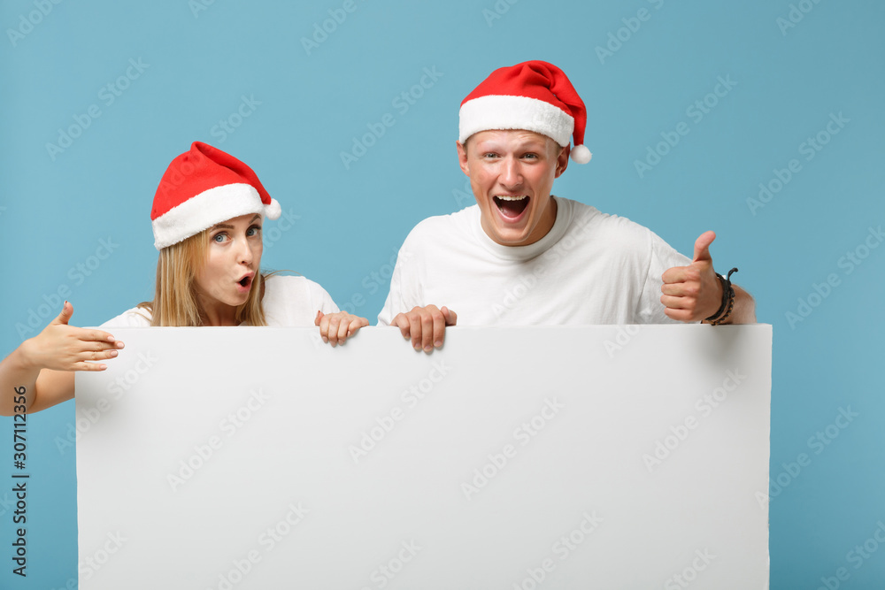 Excited young Santa couple friends guy girl in Christmas hat isolated on blue background. Happy New Year 2020 celebration holiday concept. Mock up copy space. Holding big white empty blank billboard. Excited young Santa couple friends guy girl in Christmas hat isolated on blue background. Happy New Year 2020 celebration holiday concept. Mock up copy space. Holding big white empty blank billboard.