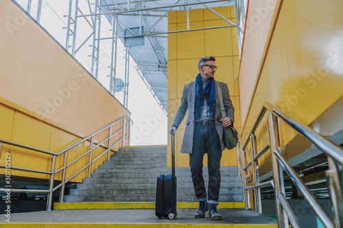 Stylish gentleman holding travel suitcase and standing on stairs